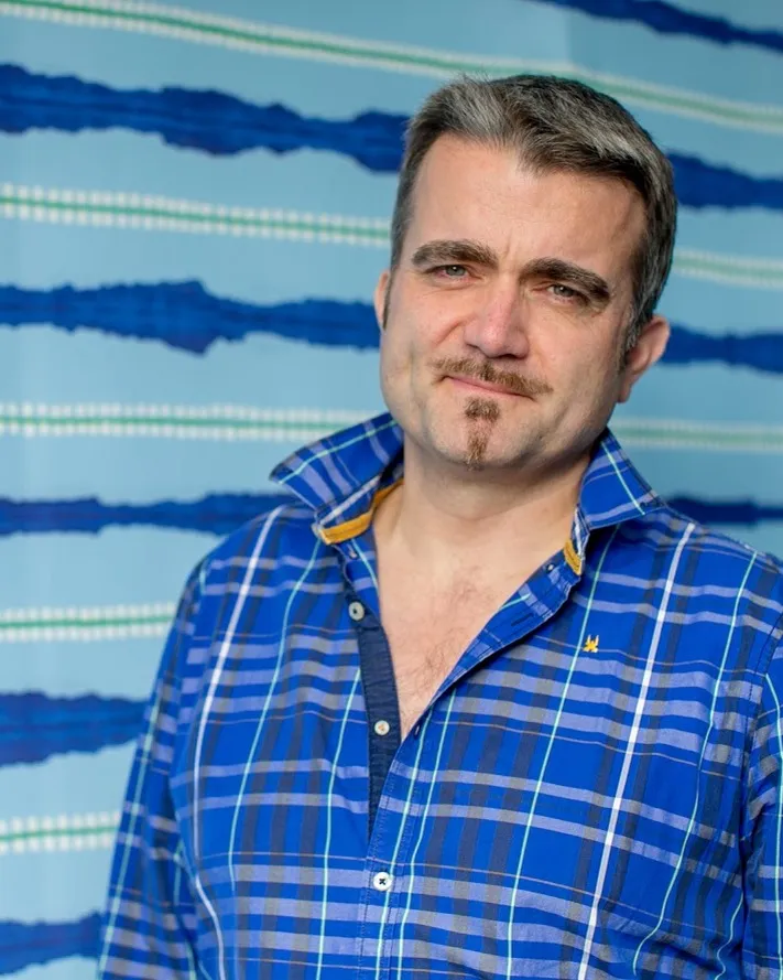 Portrait d'Olivier Collet en chemise à carreaux bleus devant un mur à rayures bleues